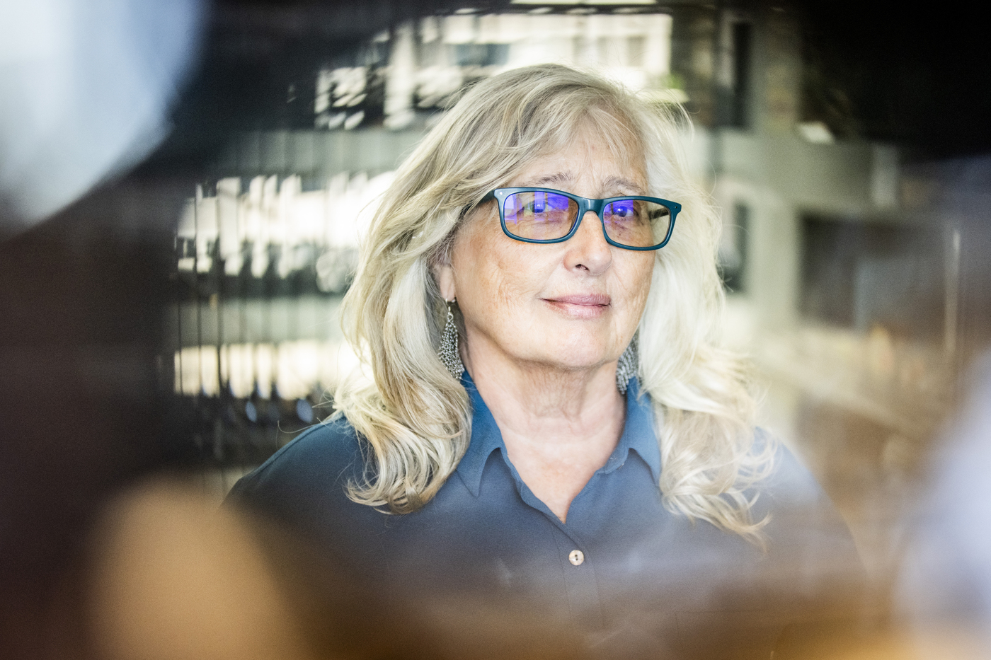 Retrato de Kathleen Kelly con un botón azul y gafas verdes en un edificio moderno. 
